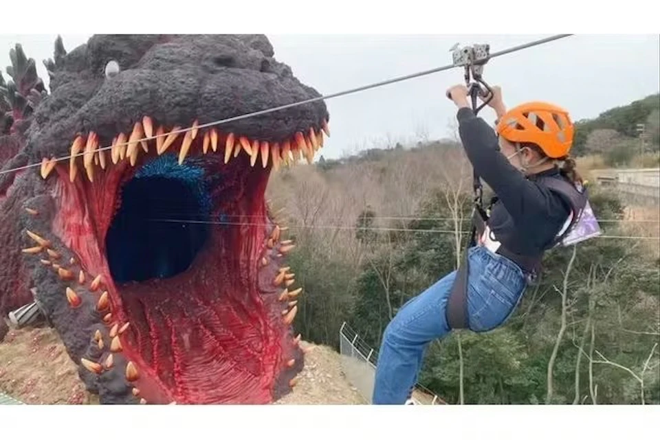 Person on a zipline approaching a large sculpture of a monster's open mouth with sharp teeth, set outdoors.