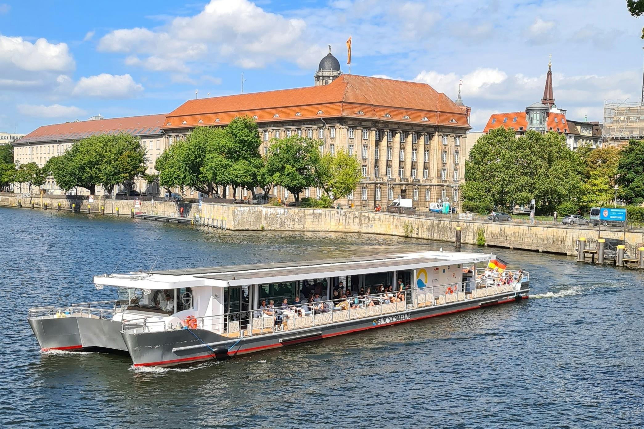 Ένα ποταμόπλοιο γεμάτο ανθρώπους ταξιδεύει σε ένα ποτάμι. Στο βάθος είναι ένα ιστορικό κτίριο με κόκκινη στέγη και δέντρα.