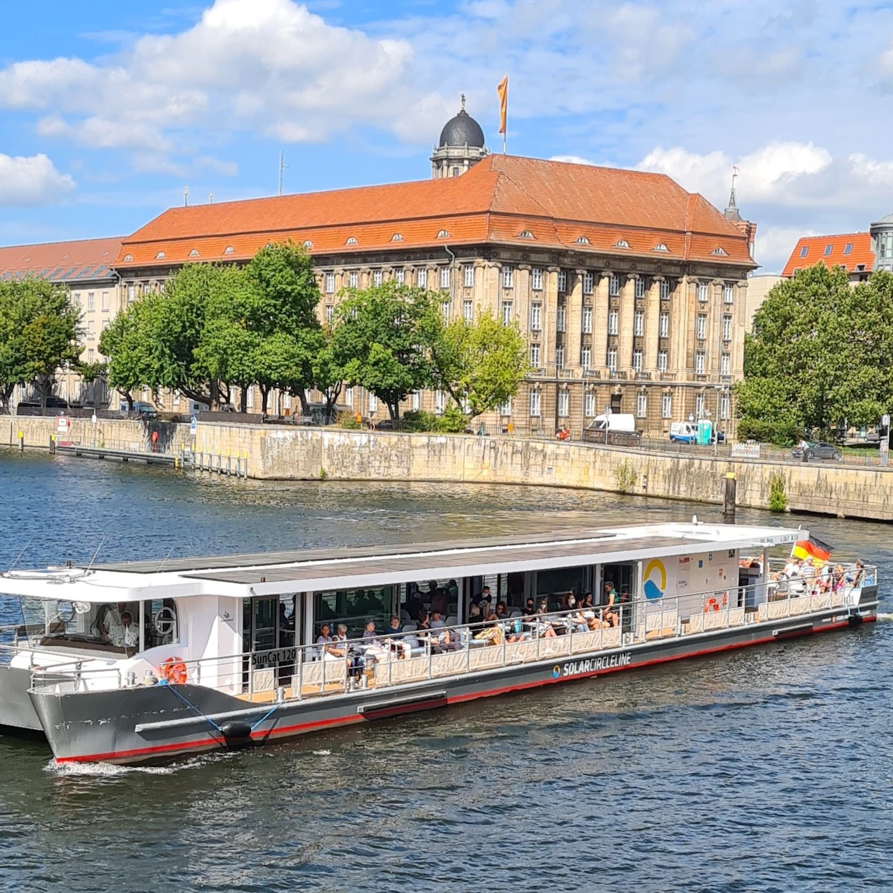 A riverboat full of people is cruising on a river; in the background is a historic building with a red roof and trees.