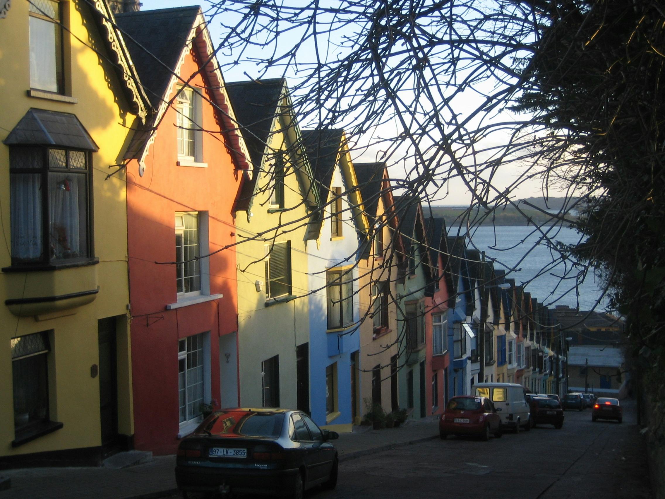 Row of colorful houses on a sloped street with cars parked alongside; water and distant land visible in the background.