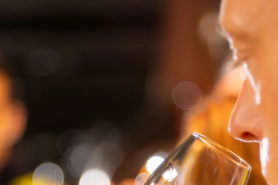 A person holding a wine glass with a golden liquid, with other people blurred in the background.