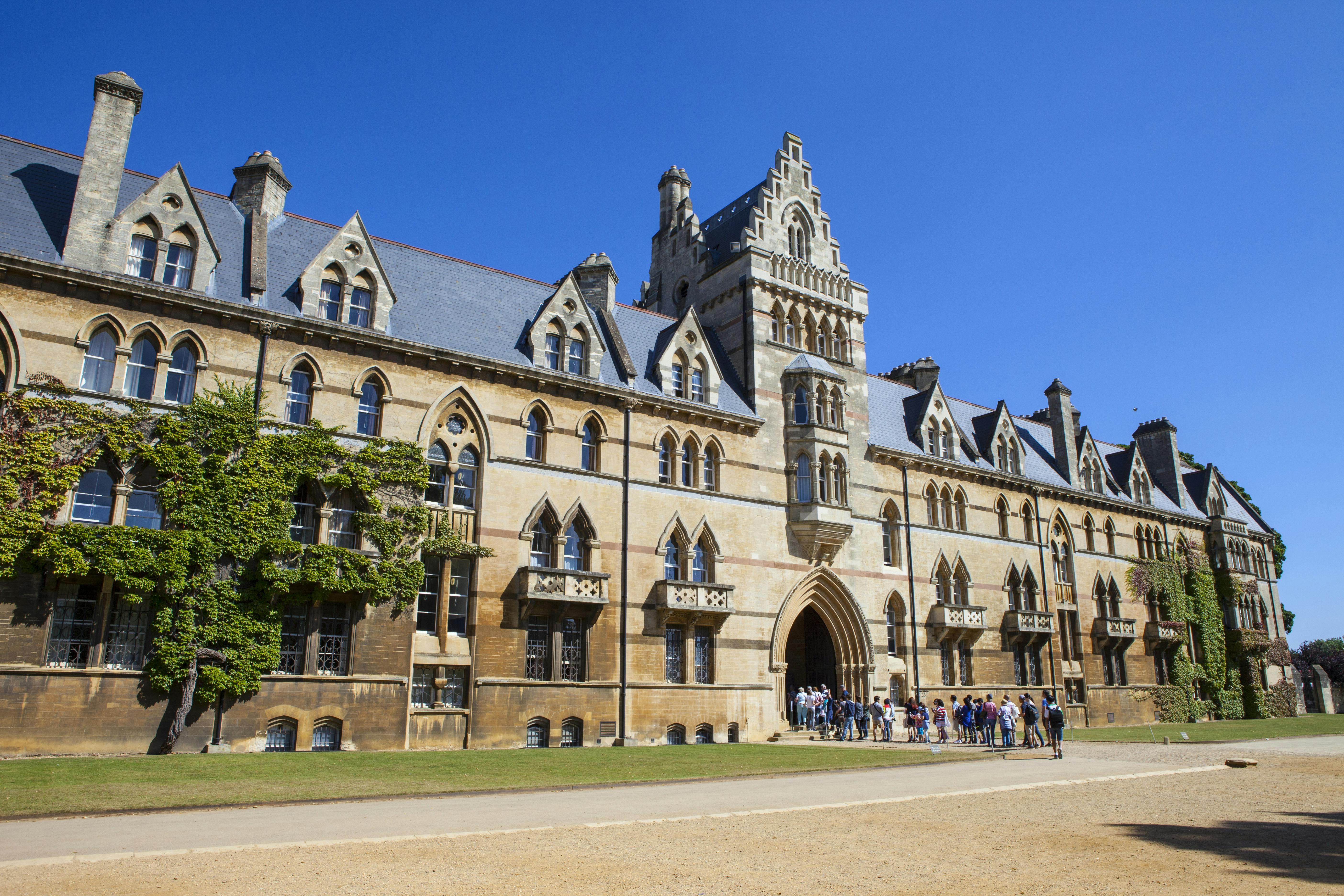 A large historic building with gothic architecture and pointed arches. A group of people is gathered near the entrance.