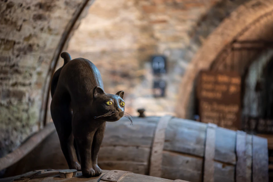 A black cat statue stands on top of a wooden barrel in a rustic, arch-bricked cellar.