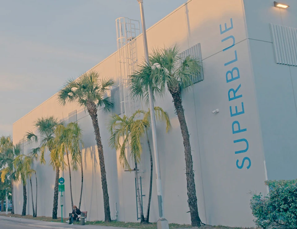 A person sits on a bench beside a large white building with "SUPERBLUE" written on it, lined with tall palm trees.