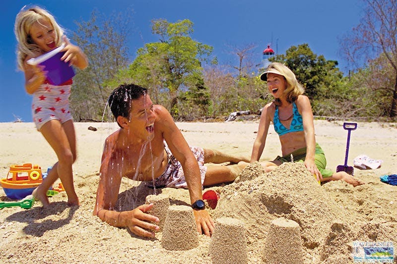 A man and woman laugh and build sandcastles on a sunny beach with trees and a red lighthouse in the background.