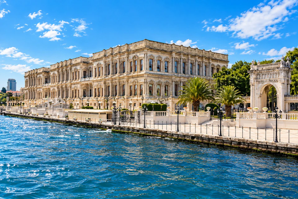 A historic ornate building with many windows, palm trees, and a waterfront under a sunny blue sky with scattered clouds.
