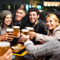 En grupp på fem vänner, som ler och håller i öl, skålar vid ett utomhusbord med mat.