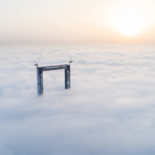 A tall structure with cranes on top emerges through thick clouds under a bright, hazy sky at sunrise.