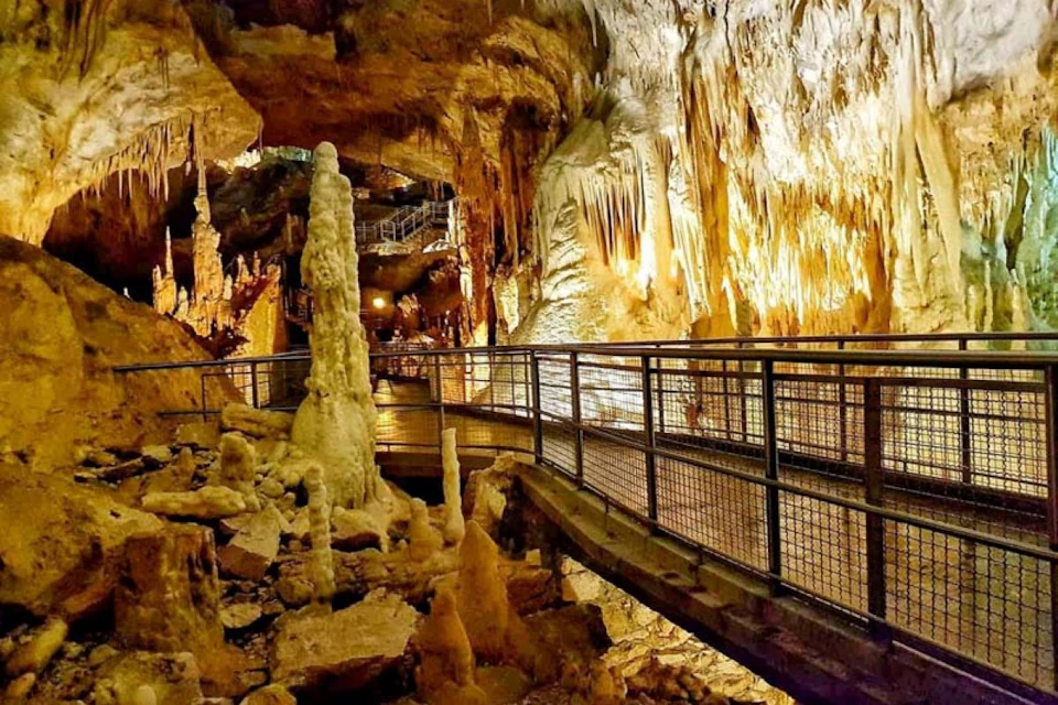 A well-lit cave interior with stalagmites, stalactites, and a metal walkway for visitors.