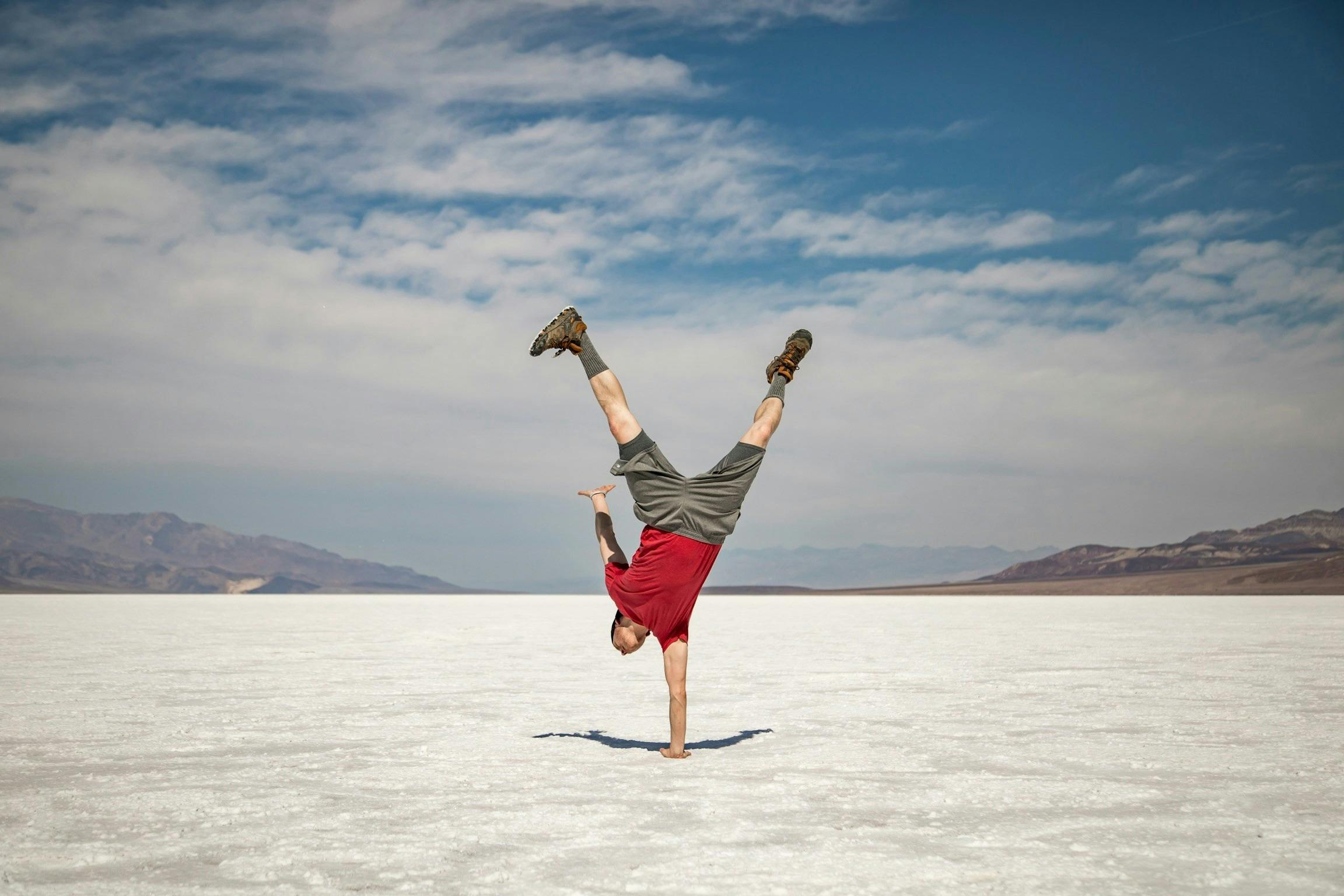 A person performs a one-handed handstand on a vast salt flat under a cloudy blue sky.