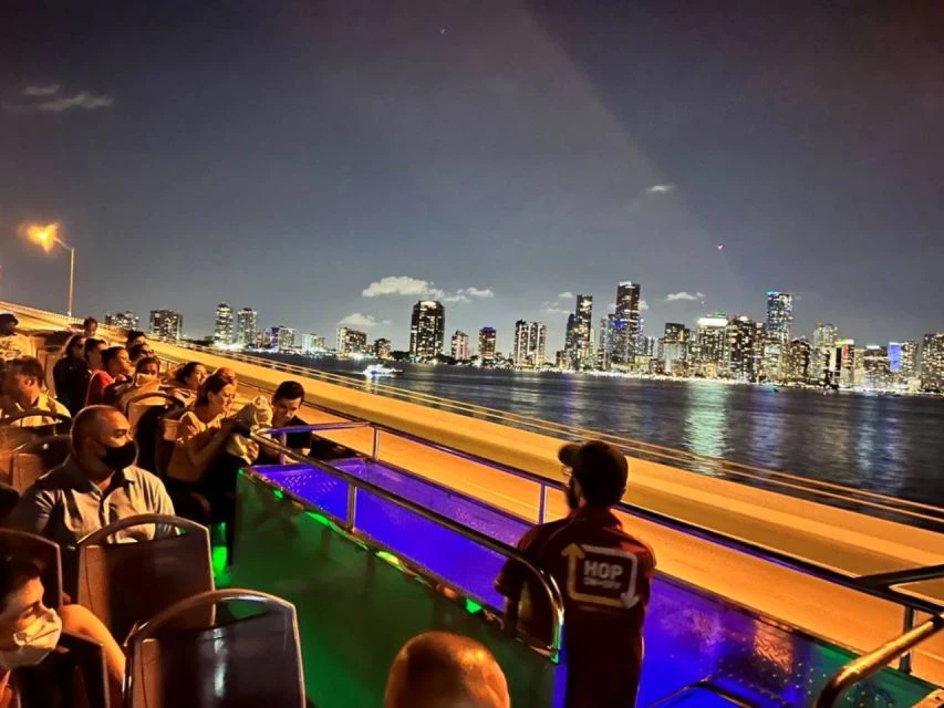 People sitting on an open-top tour bus at night, viewing a city skyline illuminated against a dark sky across a body of water.