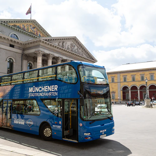 Ein blauer Doppeldeckerbus mit der Aufschrift "Münchener Stadtrundfahrten" parkt vor einem klassizistischen Säulengebäude.