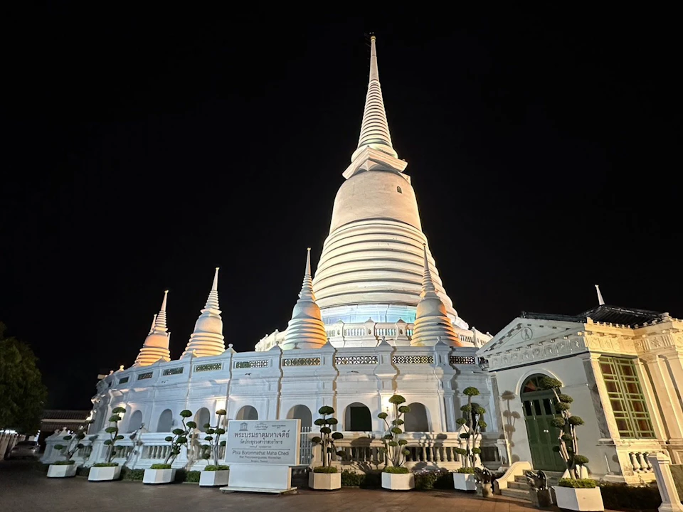 A well-lit white stupa with multiple smaller pagodas and greenery around the base, set against a dark night sky.