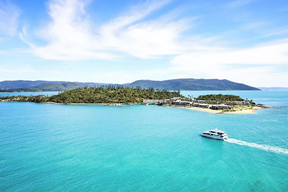 A boat sails near a tropical island with lush greenery, sandy beaches, and several buildings under a partly cloudy blue sky.