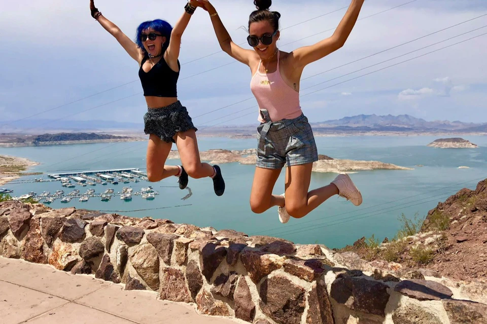 Two women are jumping in the air by a stone wall with a lake and boats in the background. Both are wearing summer outfits.
