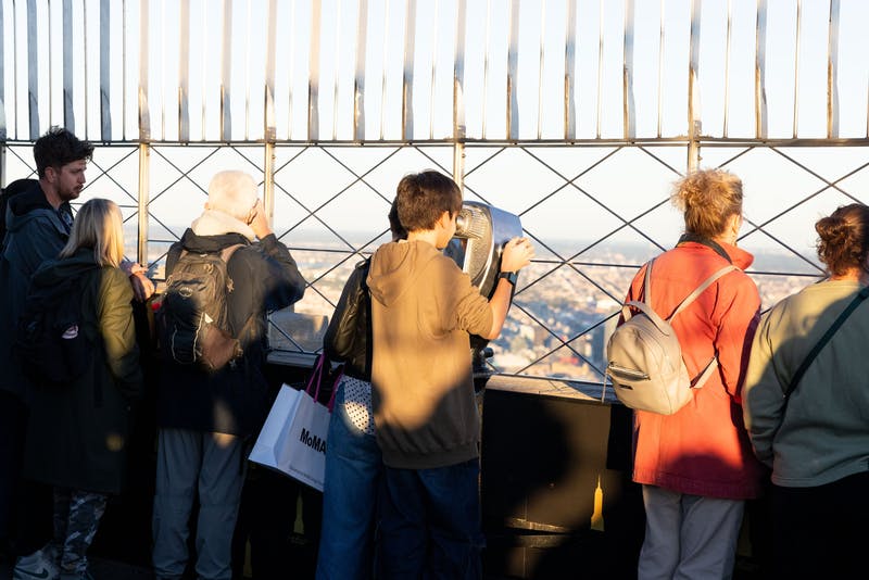 Flere mennesker står på et observationsdæk og kigger gennem søgere og over et rækværk i et bybillede.
