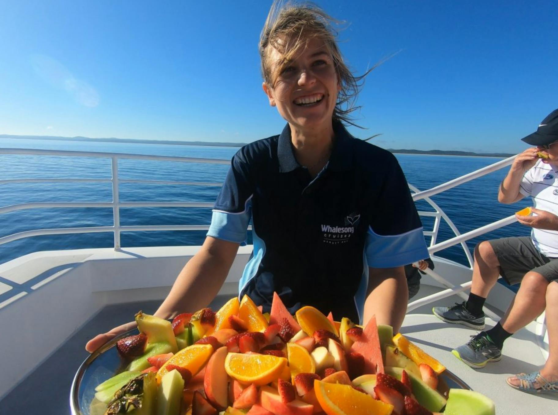 Eine lächelnde Person auf einem Boot hält einen Teller mit aufgeschnittenen Früchten unter einem strahlend blauen Himmel.