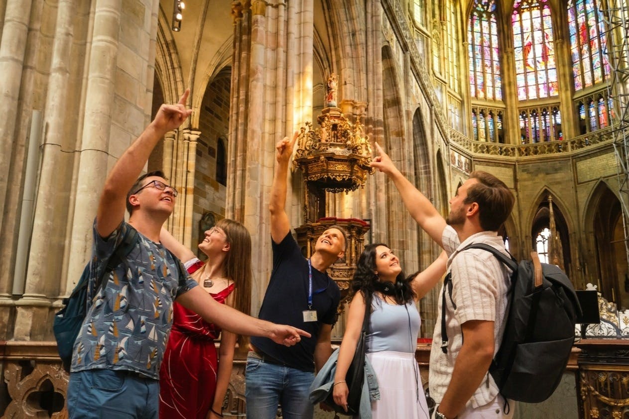 A group of people inside a ornate cathedral, pointing upwards, with tall arches and detailed carvings in the background.