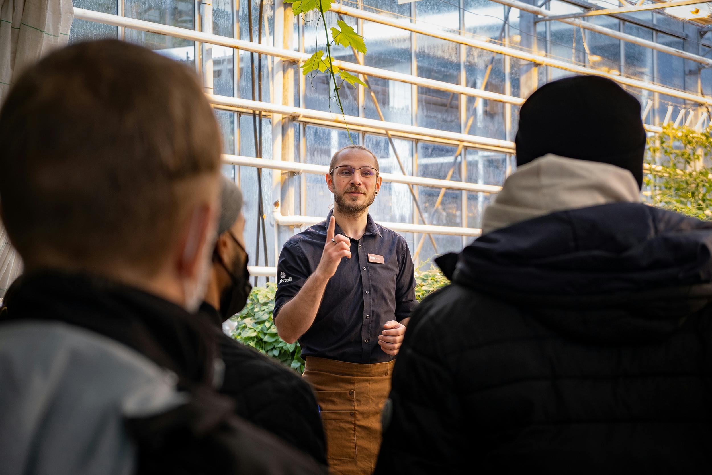 A man wearing a dark shirt and apron gestures while speaking to a group of people inside a greenhouse.