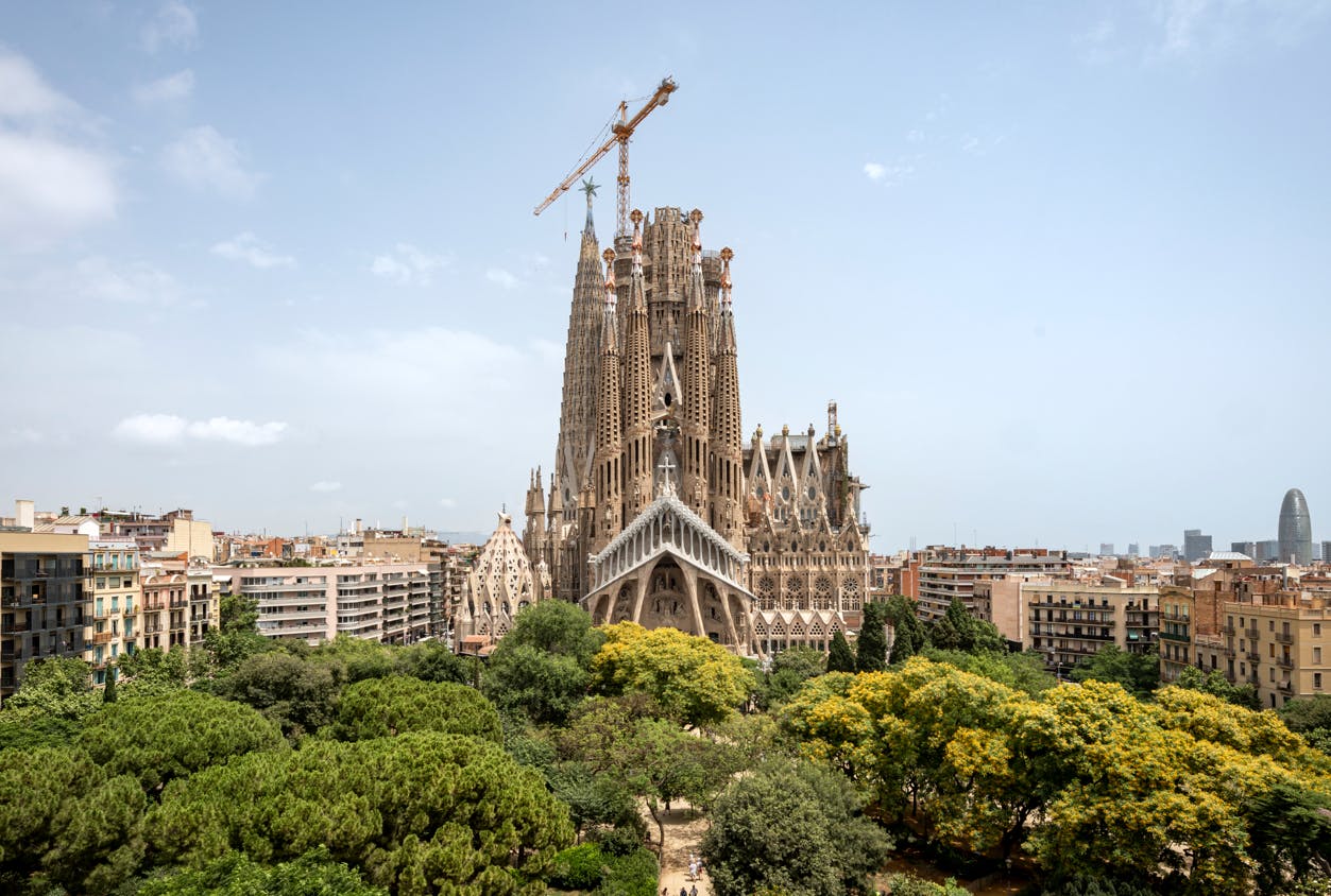 A large, ornate cathedral under construction with a crane on top, surrounded by trees and buildings under a clear sky.