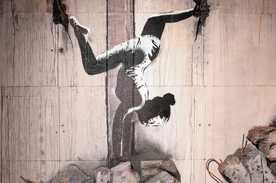 A stencil artwork of a person doing a handstand, painted on a wall with rubble and debris at the bottom.
