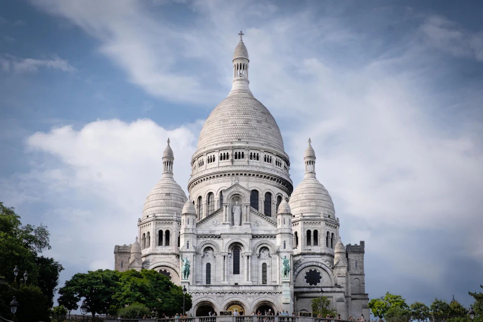A white domed basilica with ornate architecture, set against a partly cloudy blue sky and surrounded by greenery.