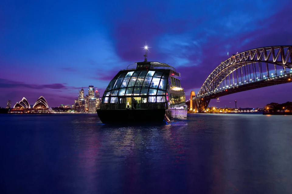 A modern glass-walled ferry illuminated at night on water with a city skyline and bridge in the background.