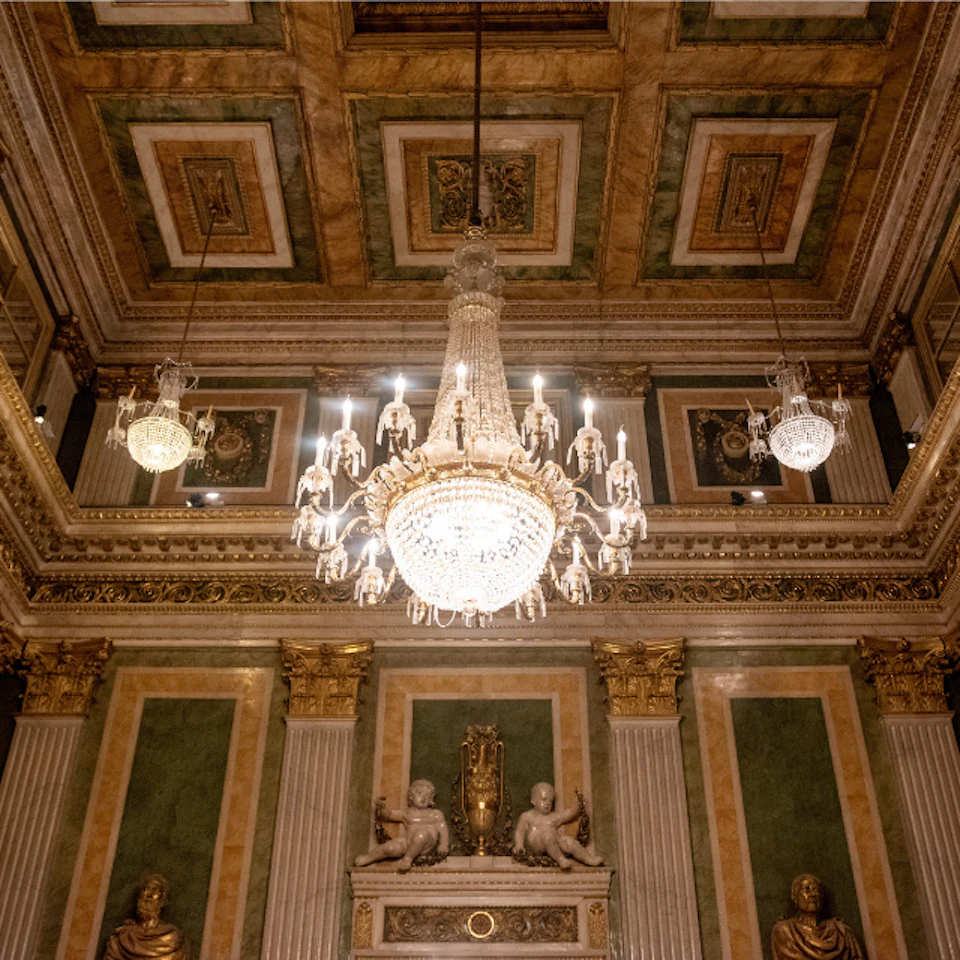 An ornate room with a grand chandelier, decorative ceiling, and classical sculptures on a wall with green panels.