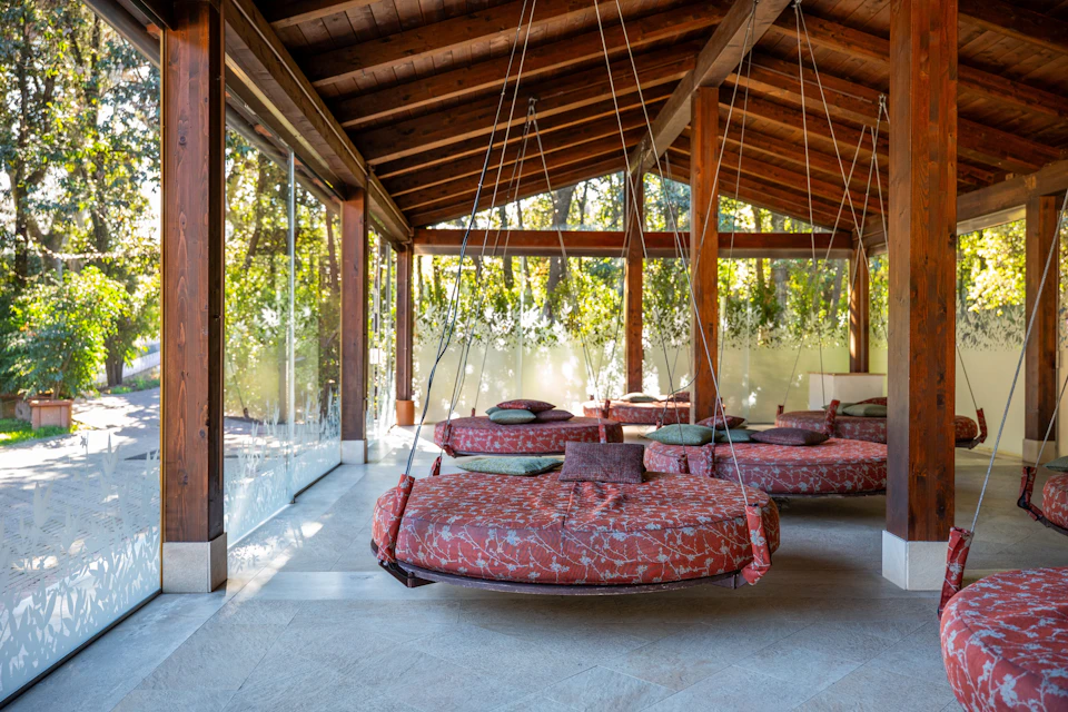 Hanging round cushioned seats with red patterned covers in a wooden-roofed space with large glass walls overlooking greenery.