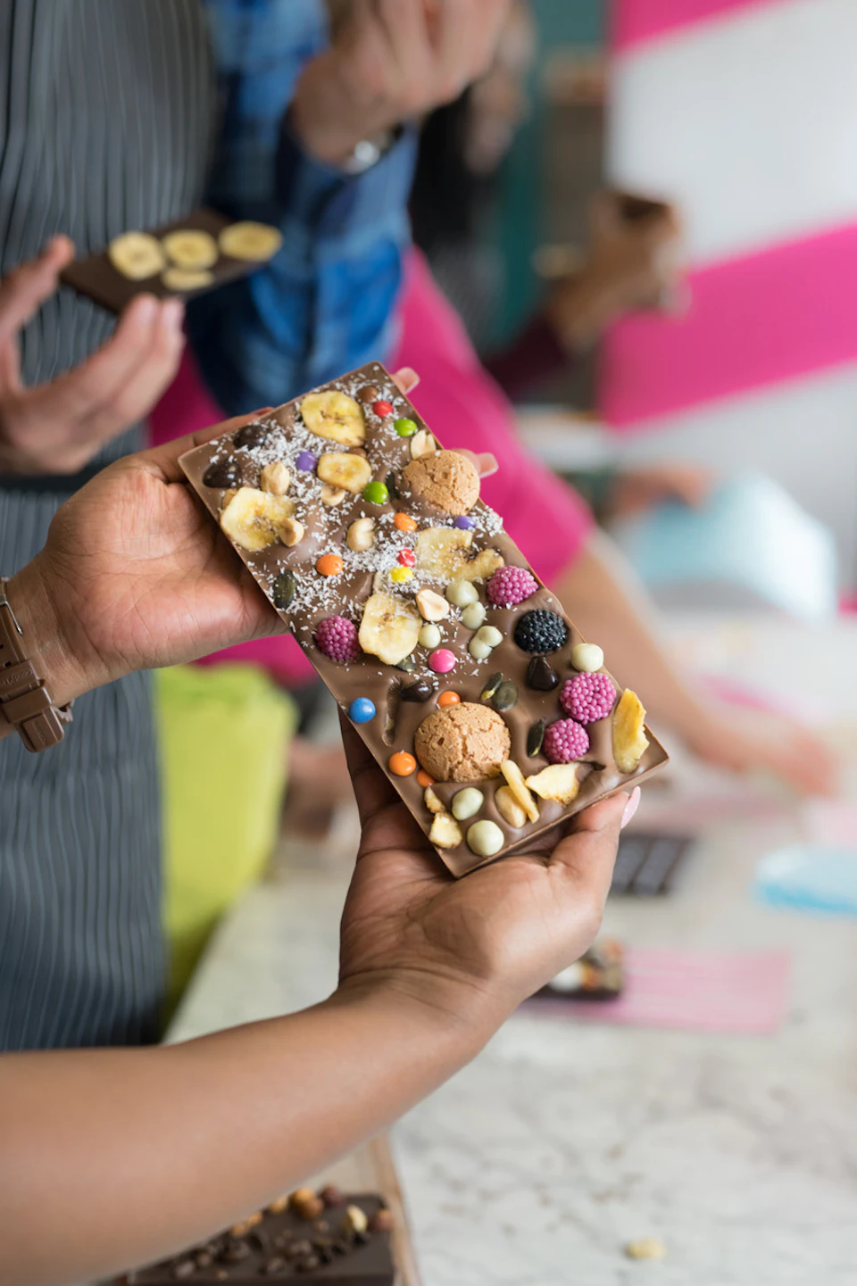 A person holding a chocolate bar covered with various toppings, including cookies, dried fruits, and colorful candies.