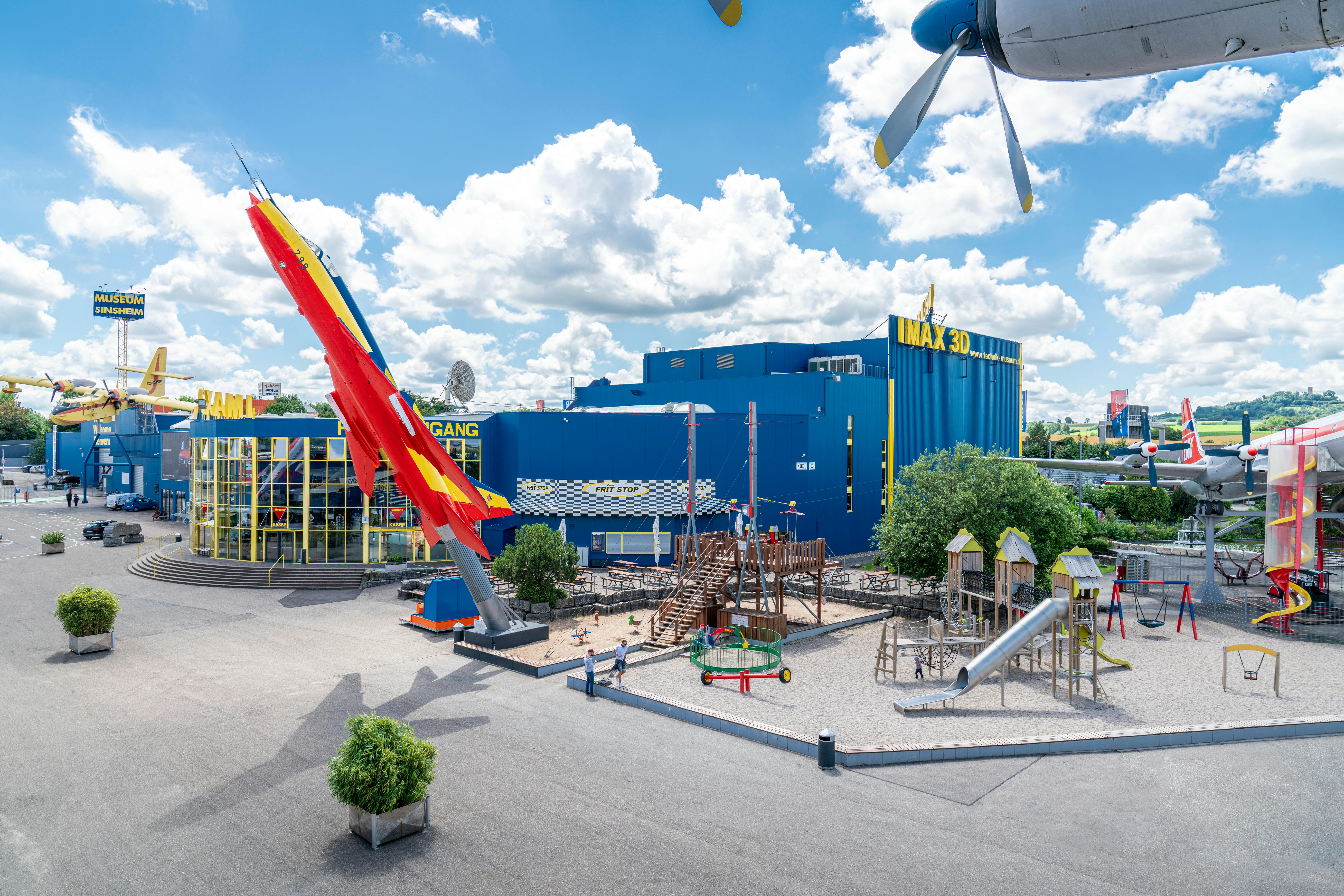 Museets entré med en färgglad flygplansskärm, IMAX 3D-teater och en närliggande lekplats under en blå himmel med moln.