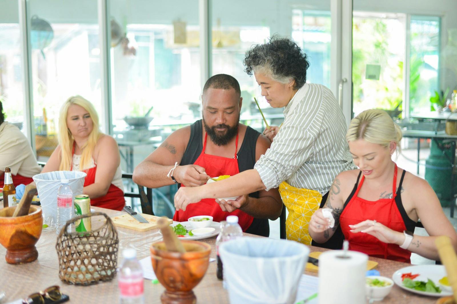四个人坐在一张桌子旁，三个人穿着红色围裙，参加一个烹饪班，由一个穿着条纹衬衫和黄色围裙的教练指导。