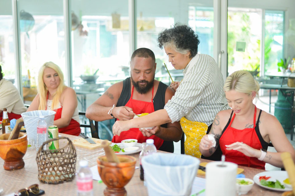 Four people at a table, three wearing red aprons, participate in a cooking class, guided by an instructor in a striped shirt and yellow apron.