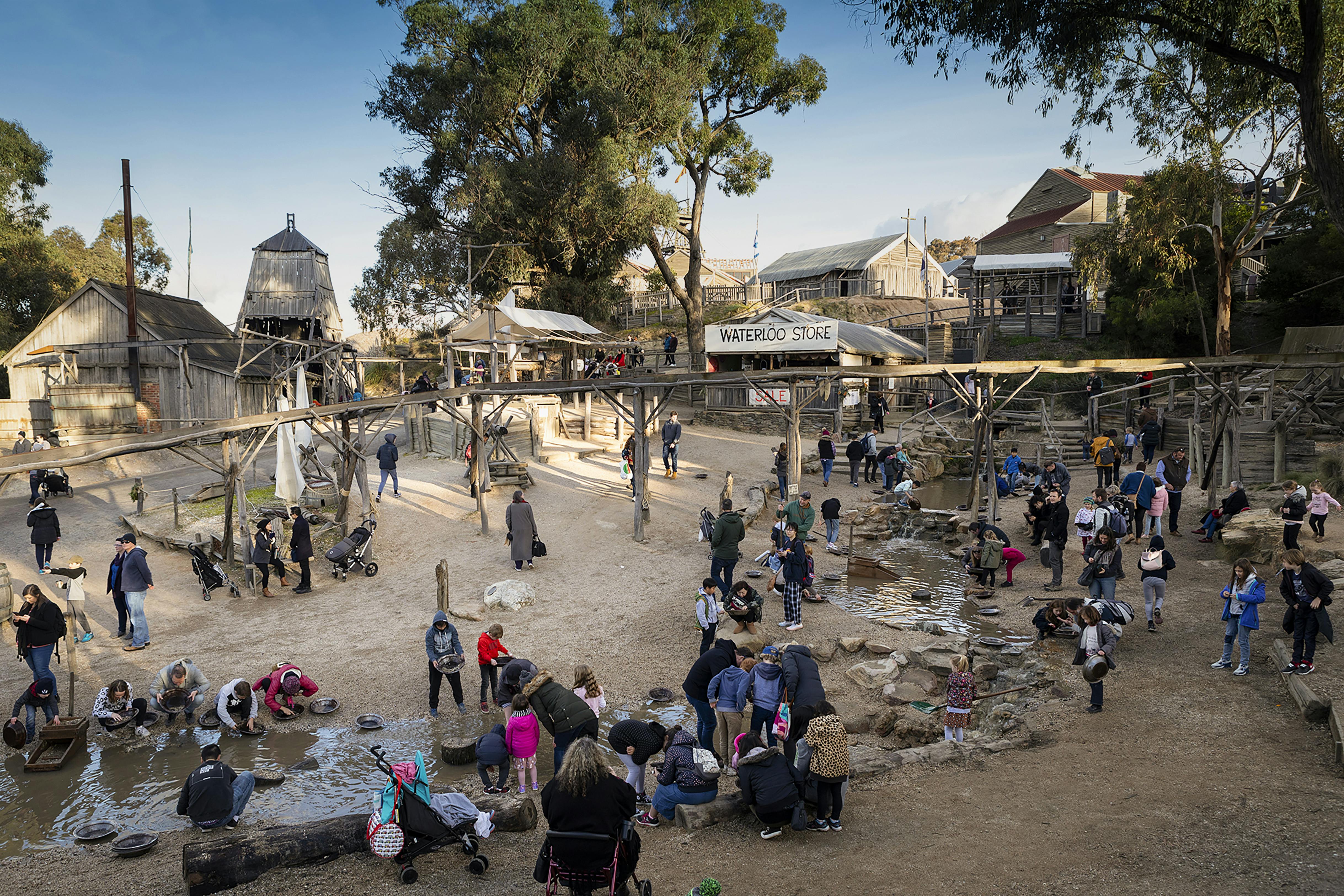Crowded outdoor historical site with people panning for gold in a shallow stream, rustic buildings, and large trees in the background.