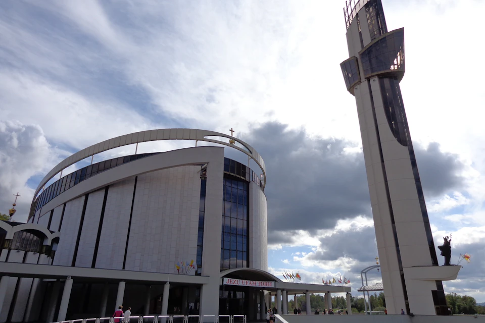 Modern church with a curved roof, cross on top, and adjacent tall tower under a cloudy sky. Some people are visible near the entrance.