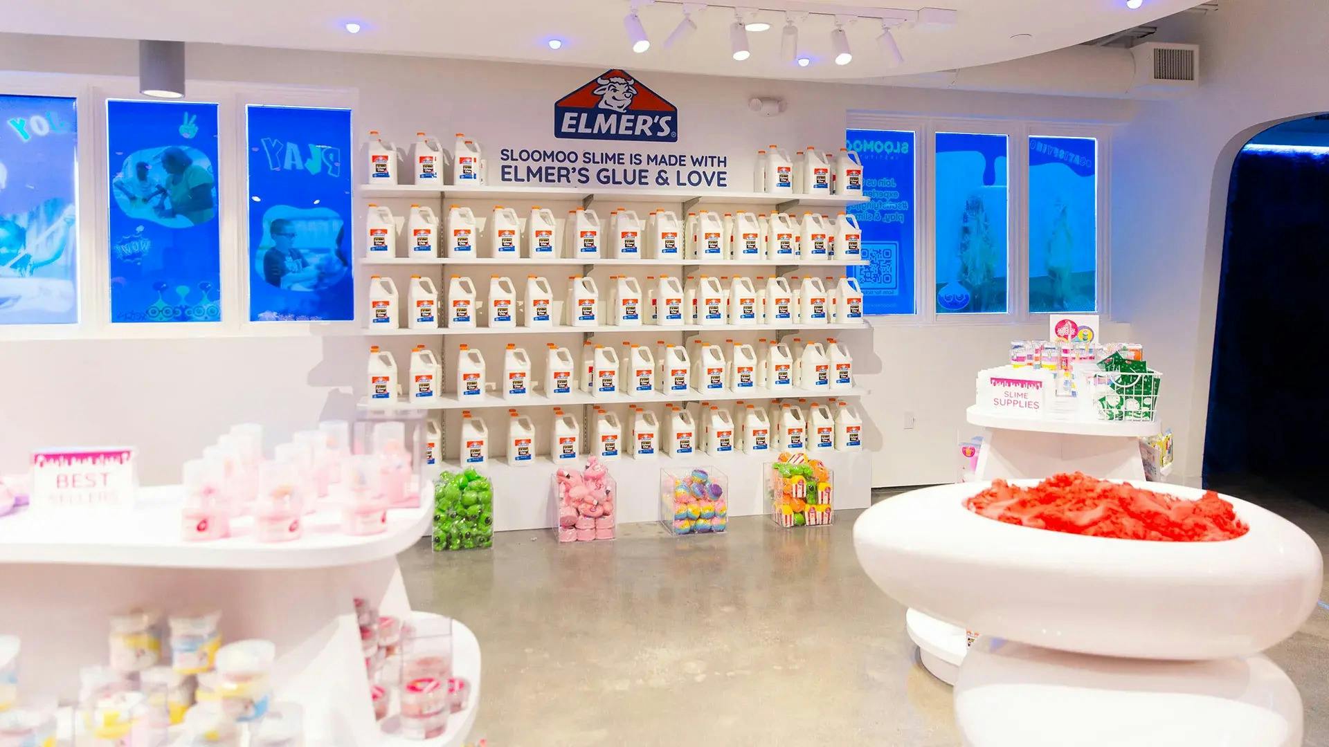 Výstava Elmerových lahví s lepidlem umístěných na policích v jasně osvětleném obchodě s různými barevnými slizovitými výrobky na bílých regálech.