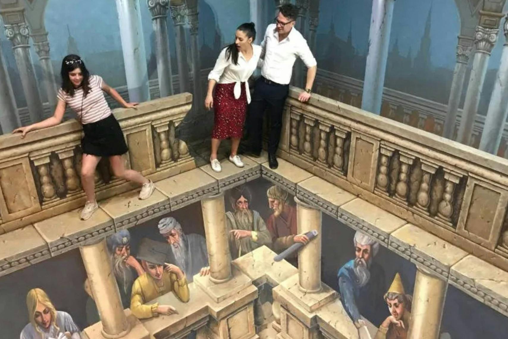Three people stand on an optical illusion artwork resembling a ledge overlooking a scene with painted historical figures.