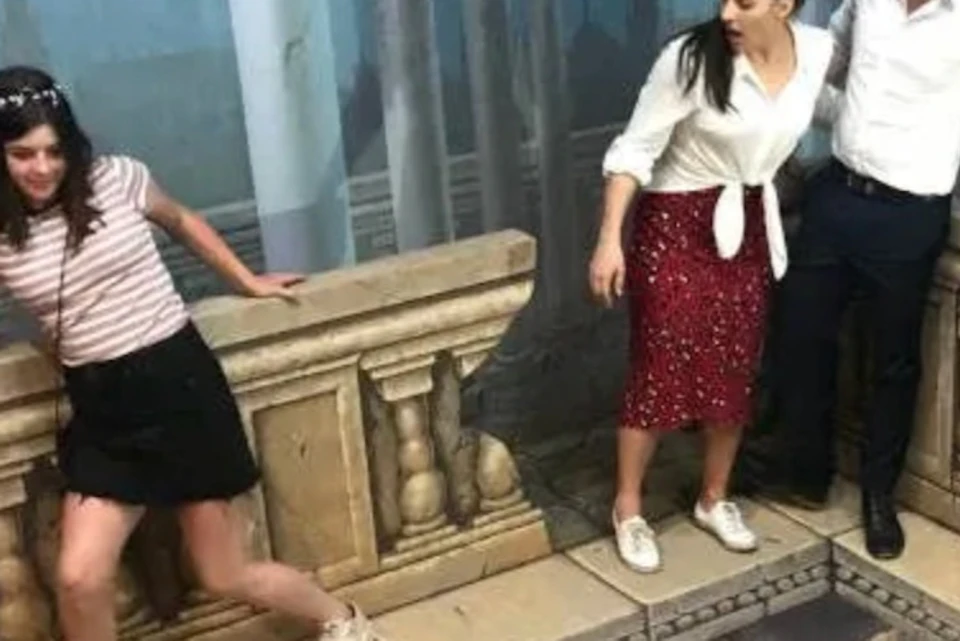 Three people stand on an optical illusion artwork resembling a ledge overlooking a scene with painted historical figures.