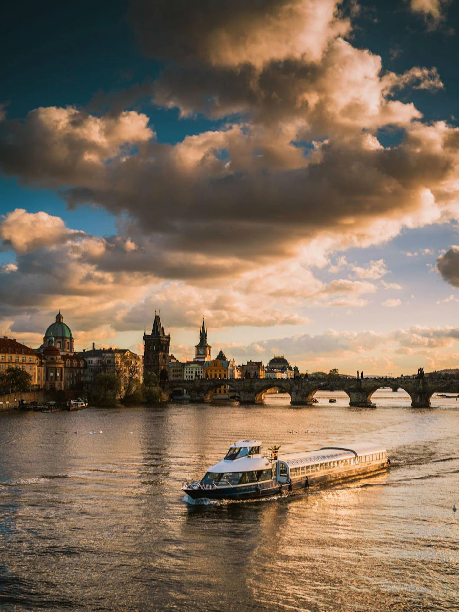A riverboat sails on a river with historic buildings and a stone bridge in the background under a cloudy sky at sunset.
