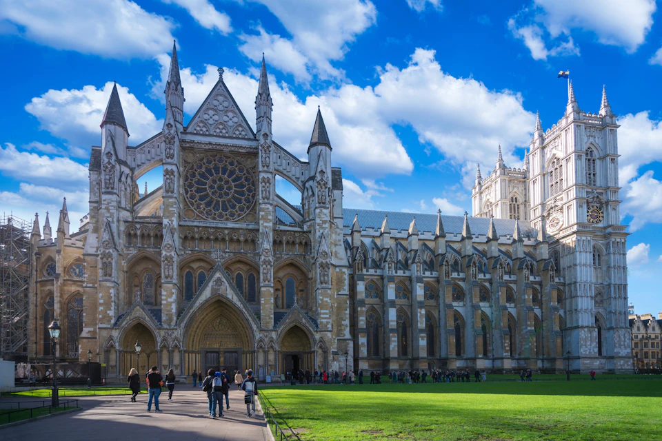 A view of a Gothic-style cathedral with ornate details, a rose window, and people walking in a grassy area under a blue sky.