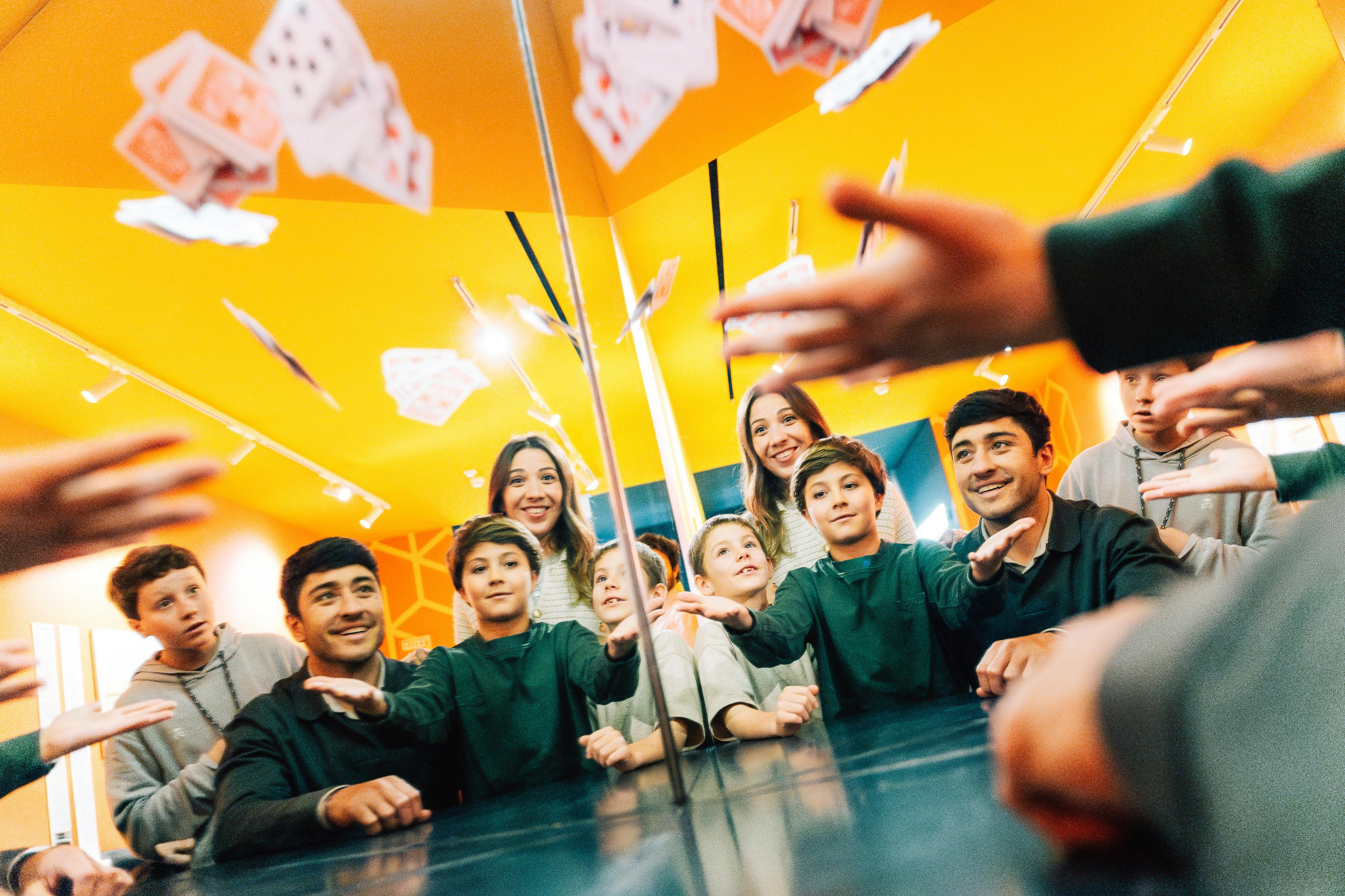 A group of smiling people in a room with mirrors and yellow walls, watching playing cards being thrown in the air.