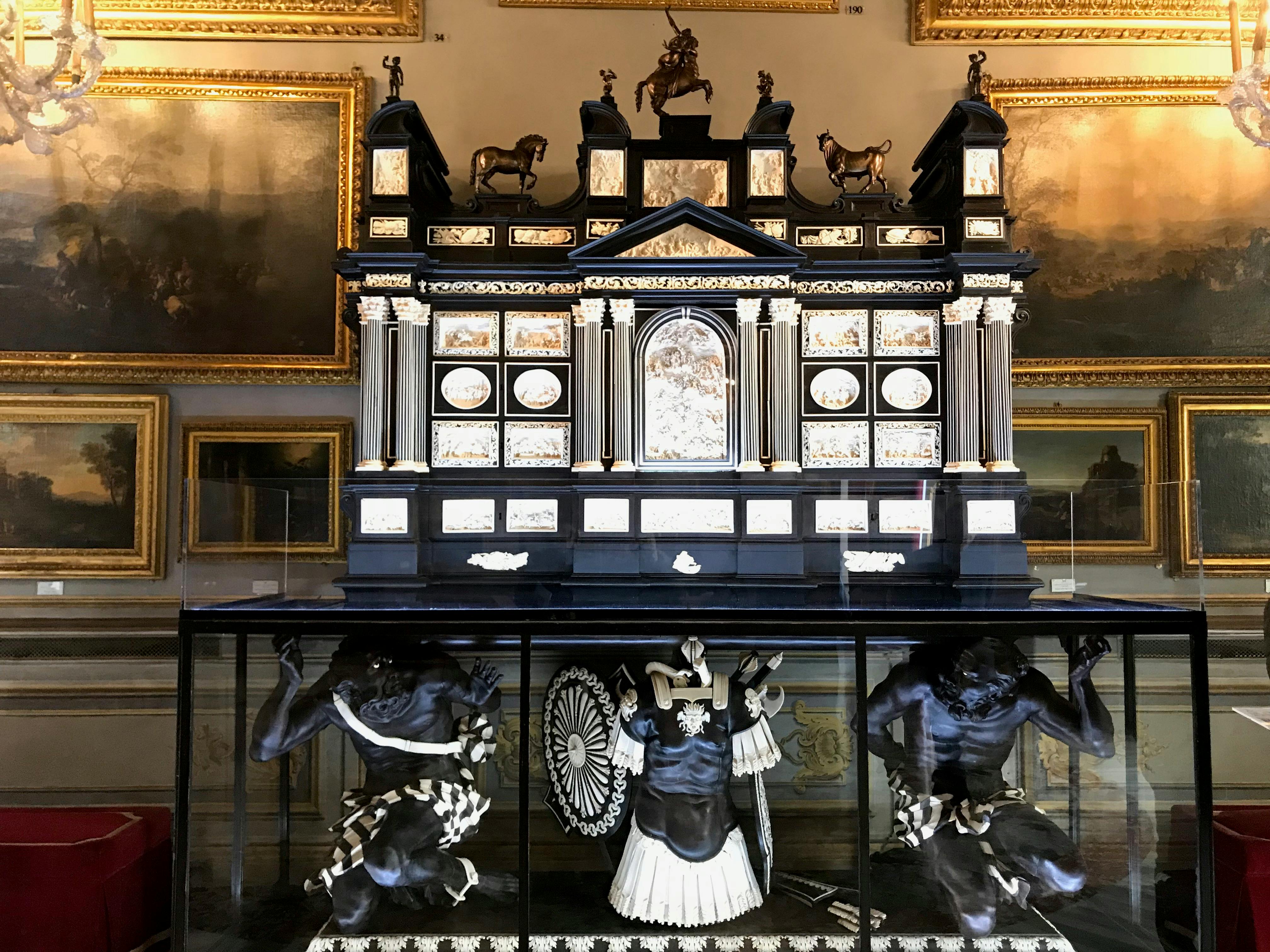 An ornate display features a detailed structure with columns and statues of animals on top, and figures beneath, encased in glass.