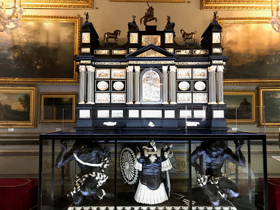An ornate display features a detailed structure with columns and statues of animals on top, and figures beneath, encased in glass.