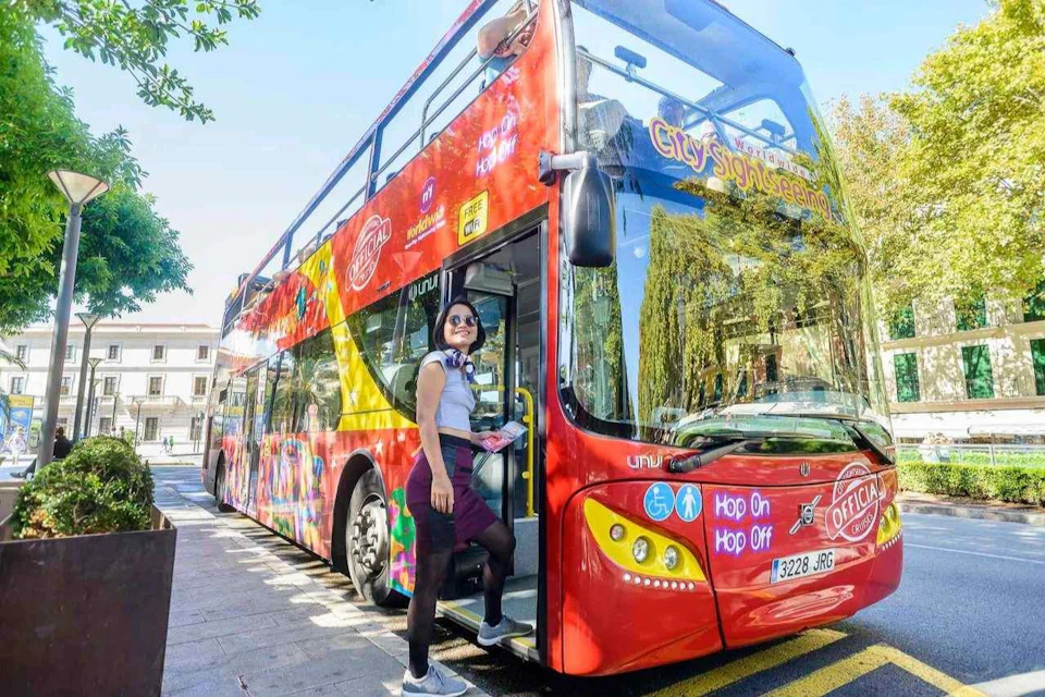 A woman stands at the entrance of a red double-decker sightseeing bus, holding a map and smiling.