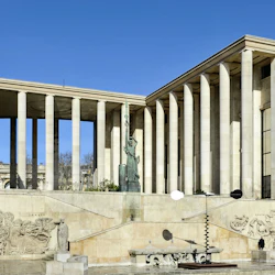 Stor byggnad med höga pelare, utsmyckade stenhuggningar på väggar och en central staty som håller en fackla under en klarblå himmel.