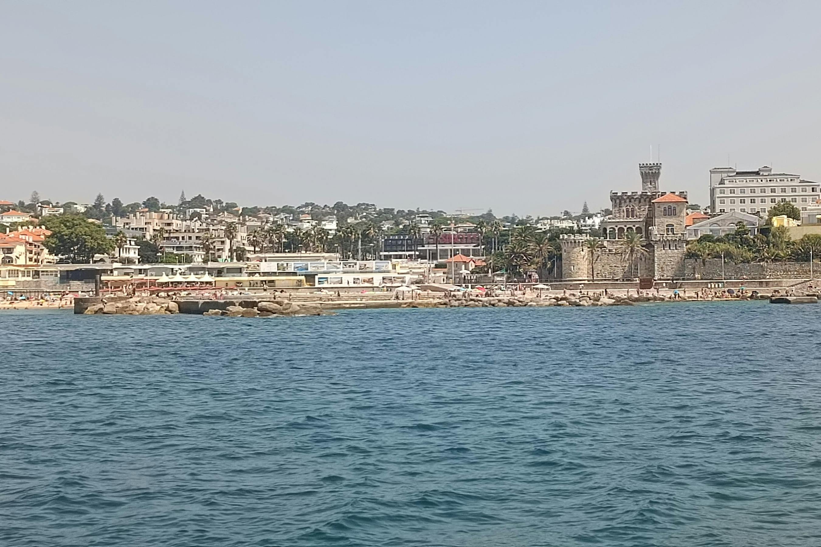 Μια παραθαλάσσια πόλη με κτίρια, έναν ιστορικό πέτρινο πύργο και μια βραχώδη ακτογραμμή, ορατή από ένα υδάτινο σώμα.