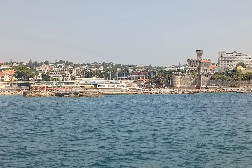 A coastal town with buildings, a historic stone tower, and a rocky shoreline, viewed from across a body of water.