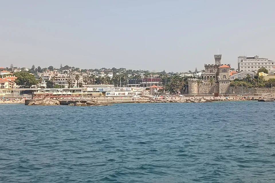 A coastal town with buildings, a historic stone tower, and a rocky shoreline, viewed from across a body of water.