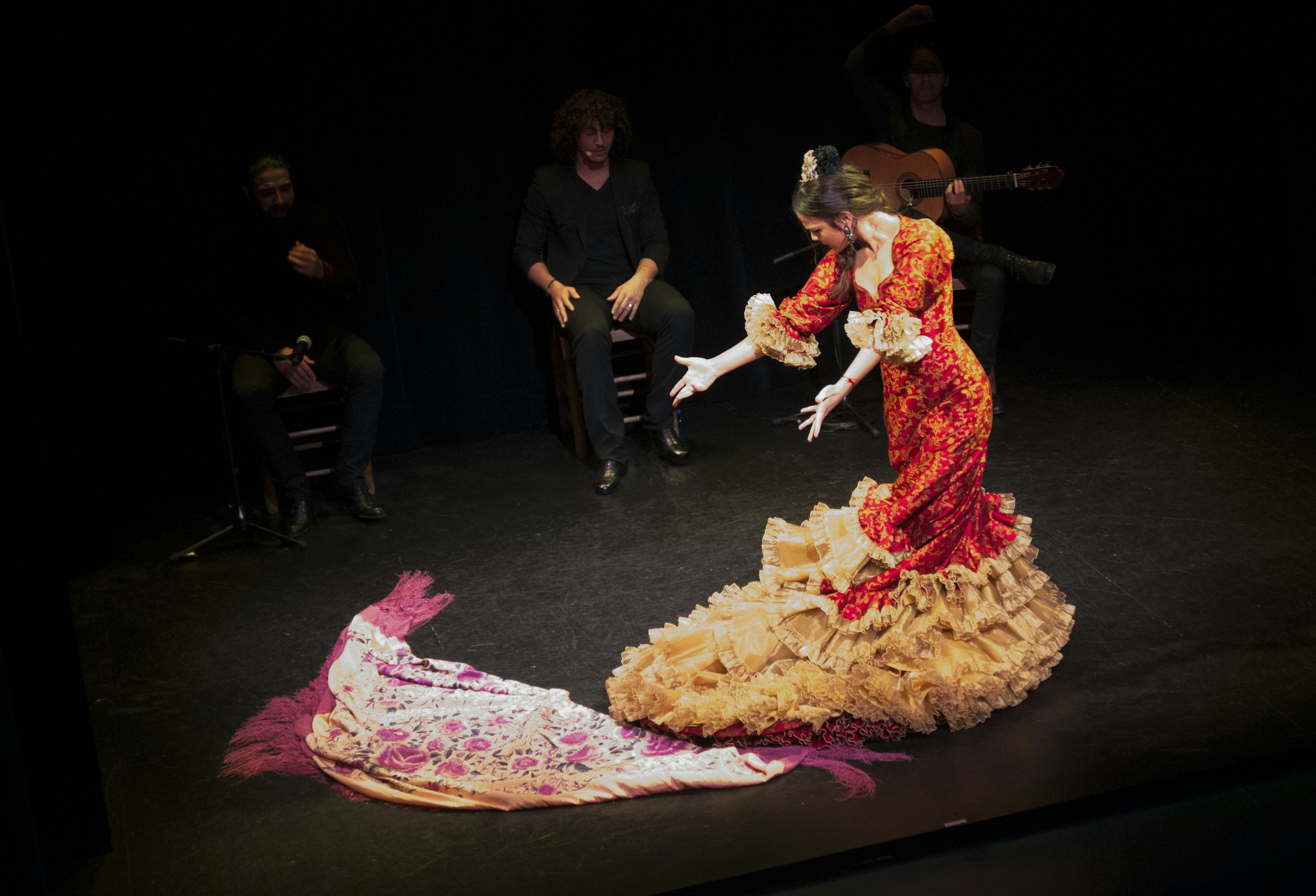 Ballarina de flamenc amb un vestit vermell i daurat amb un mantó rosa que actua a l'escenari amb músics asseguts al fons.