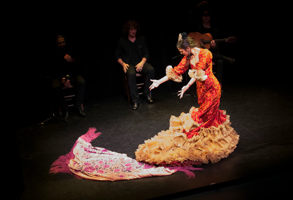 Flamenco dancer in a red and gold dress with a pink shawl performs on stage with seated musicians in the background.